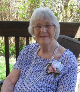 A picture of a grey haired, older, smiling lady sitting on a chair