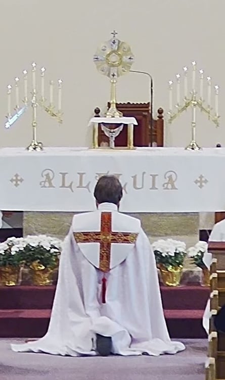 Priest kneeling in front of Monstrance, wearing white cope