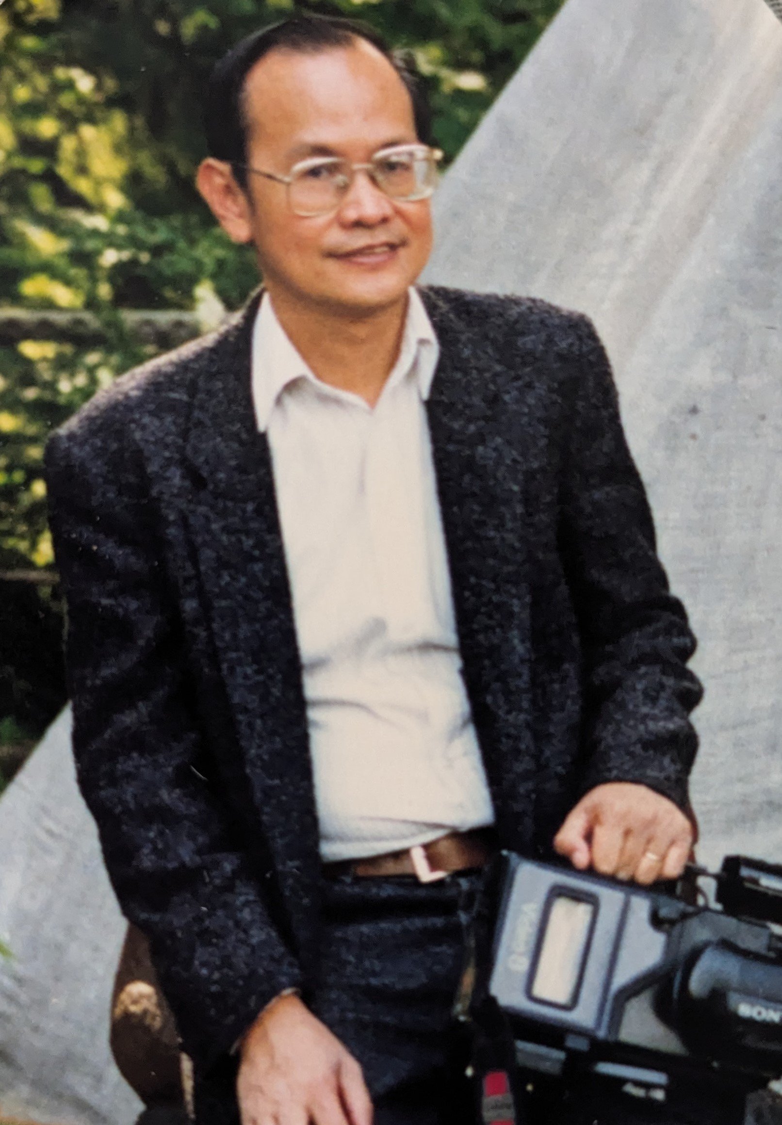 A smiling Asian man with glasses holding a video camera