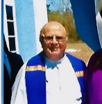An older, bald, slightly chubby priest wearing a white alb and purple stole.  He is smiling gently.